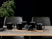 Fünf matte schwarze Keramikbecher auf Holzbrett auf einem Holztisch, eine Tasse liegt seitlich, frontal aus leicht erhöhter Perspektive vor dunklem Hintergrund