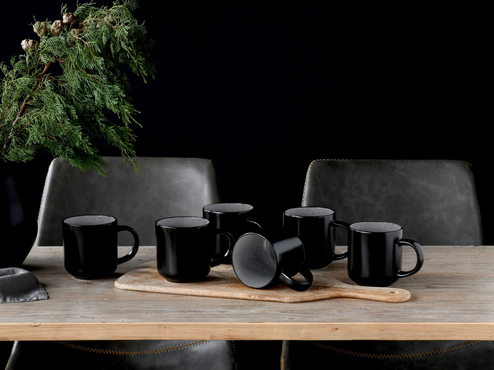 Fünf matte schwarze Keramikbecher auf Holzbrett auf einem Holztisch, eine Tasse liegt seitlich, frontal aus leicht erhöhter Perspektive vor dunklem Hintergrund