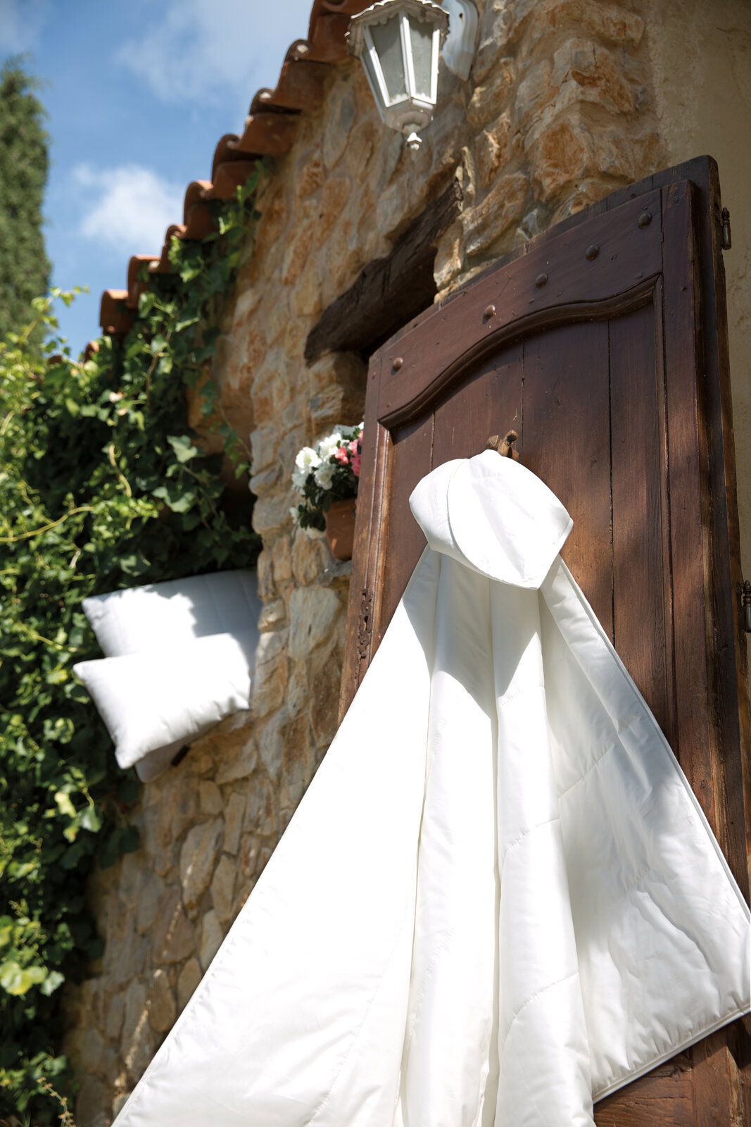 Weiße Sommerbettdecke hängt an einer Holztür vor einer Steinwand mit Efeu und Blumen, seitliche Perspektive.