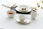 Weißes Porzellan-Geschirrset mit dezentem Rillendekor, bestehend aus Tellern, Schalen, Tasse mit Untertasse und Becher, in schräger Vorderansicht auf einem hell gedeckten Tisch arrangiert