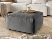 Grauer Hocker mit gerippter Textur, von schräg oben fotografiert, auf einem Holzboden vor einem cremefarbenen Sofa platziert.