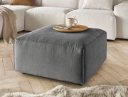 Grauer Hocker mit gerippter Textur, von schräg oben fotografiert, auf einem Holzboden vor einem cremefarbenen Sofa platziert.