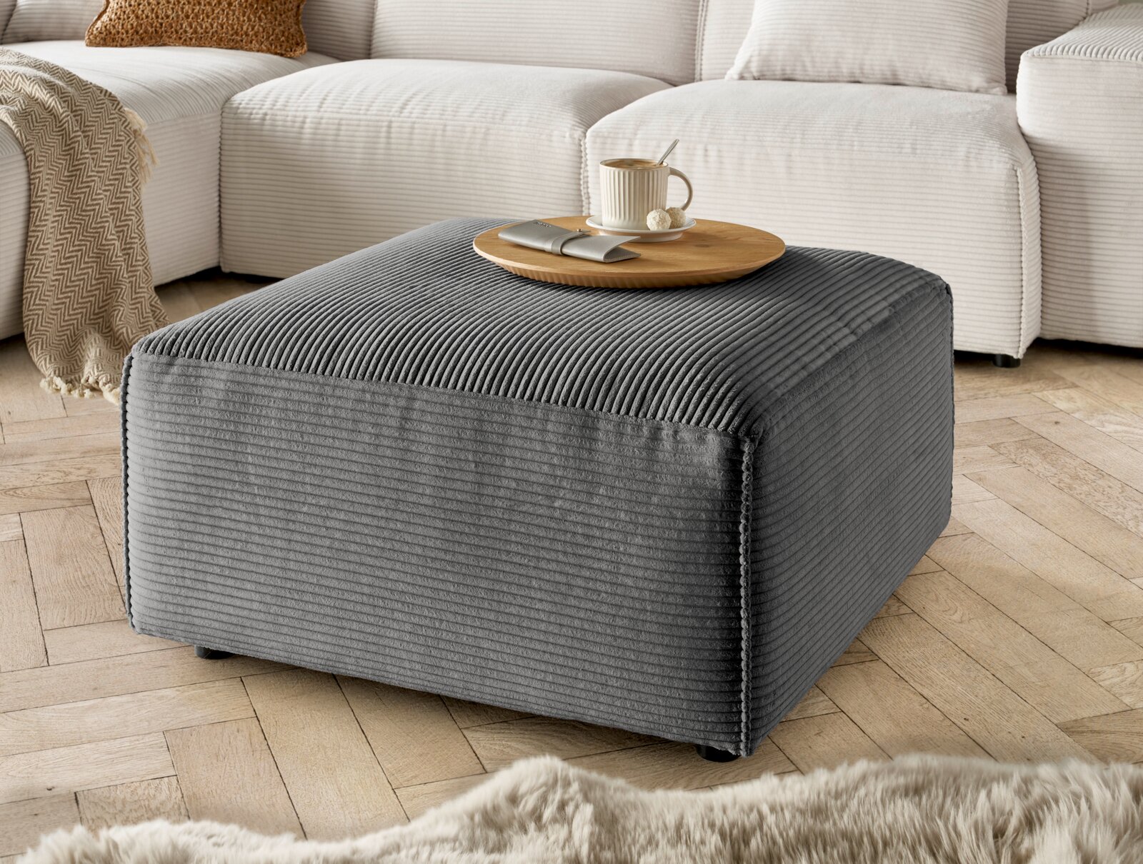 Grauer Hocker mit gerippter Textur, von schräg oben fotografiert, auf einem Holzboden vor einem cremefarbenen Sofa platziert.