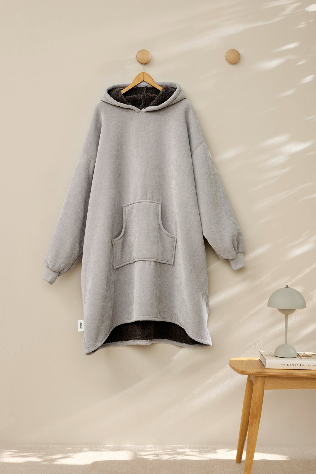 Grauer Lounge Hoodie mit Kapuze und Kängurutasche, frontal an einem Haken hängend, mit weichem Innenfutter.