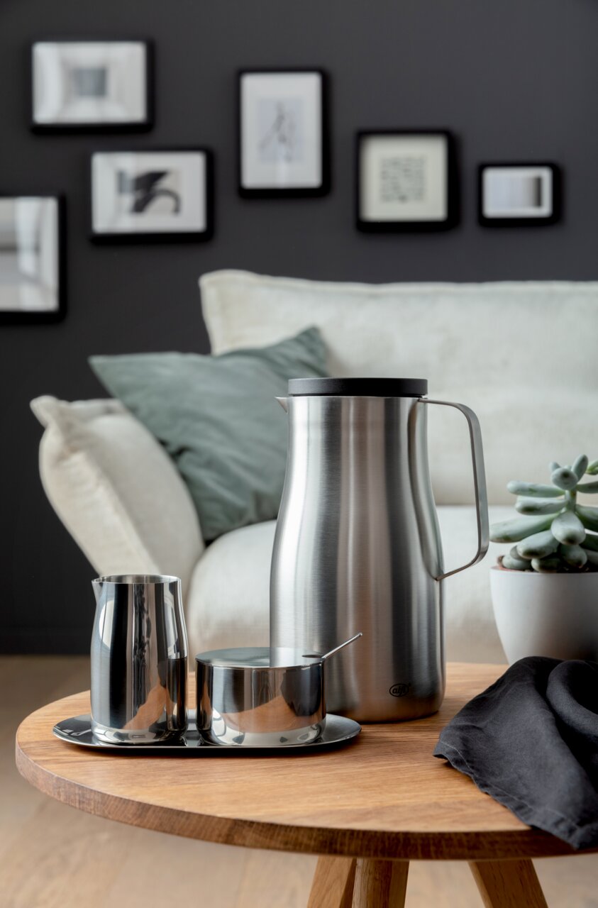 Alfi Milch & Zucker Set Elegantes Milch- und Zucker-Set aus Edelstahl auf einem Holztisch, mit einer modernen Kanne, einem Milchkännchen und einer Zuckerdose mit Löffel, im Hintergrund ein gemütliches Wohnzimmer. Perspektive: seitlich.