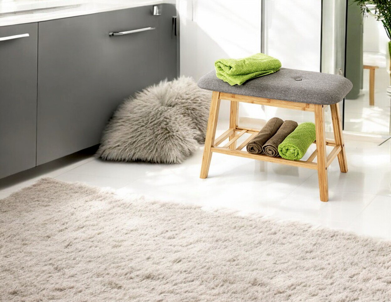 Badezimmer-Hocker/Bank aus hellem Holz mit grauer gepolsterter Sitzfläche und Ablage mit gerollten Handtüchern, aufgenommen in schräger Frontansicht im hellen Badezimmer.