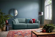 Ein modernes Funktionssofa in einem stilvoll eingerichteten Wohnzimmer, frontal betrachtet. Das Sofa ist in einem sanften Grünton gehalten und mit bunten Kissen dekoriert. Im Hintergrund sind eine blaue Wand, ein runder Spiegel und ein großes Fenster zu s