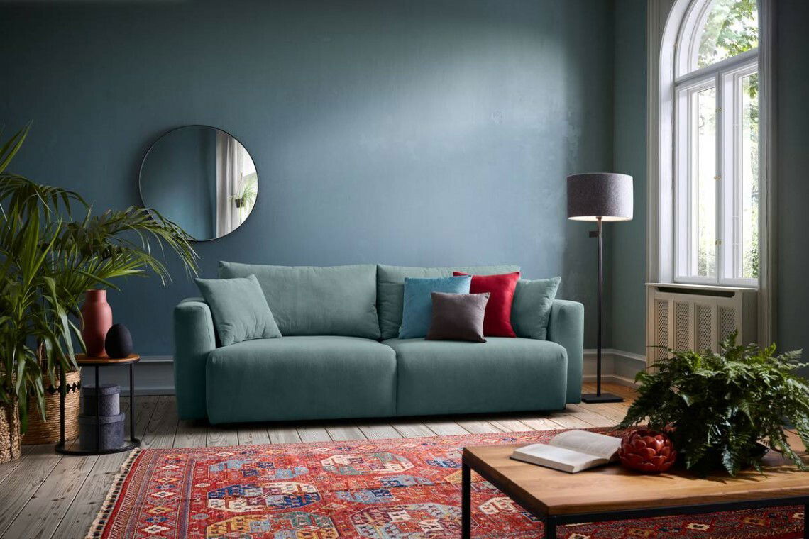 Ein modernes Funktionssofa in einem stilvoll eingerichteten Wohnzimmer, frontal betrachtet. Das Sofa ist in einem sanften Grünton gehalten und mit bunten Kissen dekoriert. Im Hintergrund sind eine blaue Wand, ein runder Spiegel und ein großes Fenster zu s