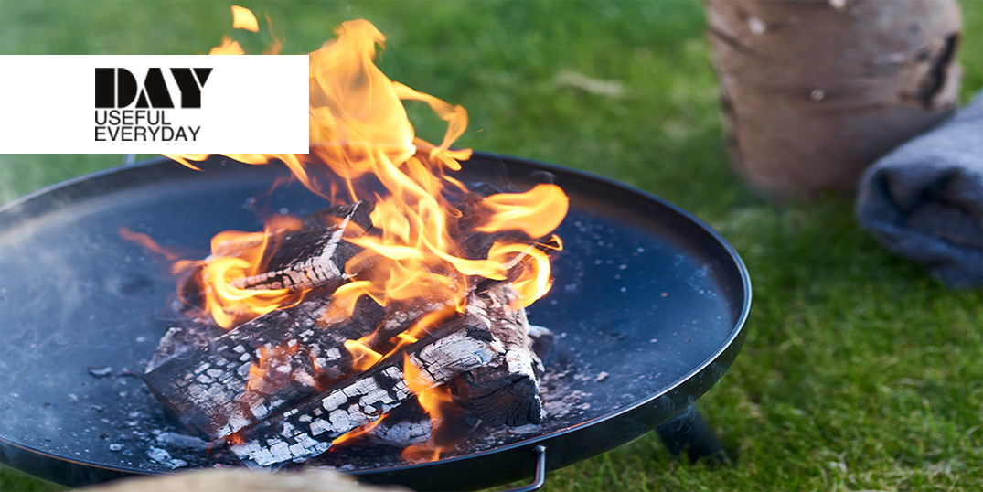Auf einer Wiese steht eine schwarzen Feuergrube mit loderndem Lagerfeuer.