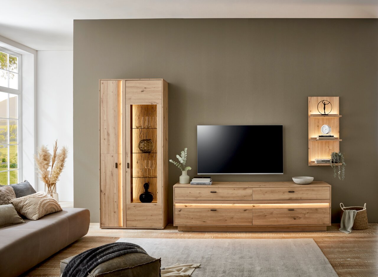 Moderne Wandkombination aus Holz in einem Wohnzimmer, bestehend aus einem hohen Schrank mit Glaselementen, einem TV-Board und einem Wandregal, seitliche Perspektive.