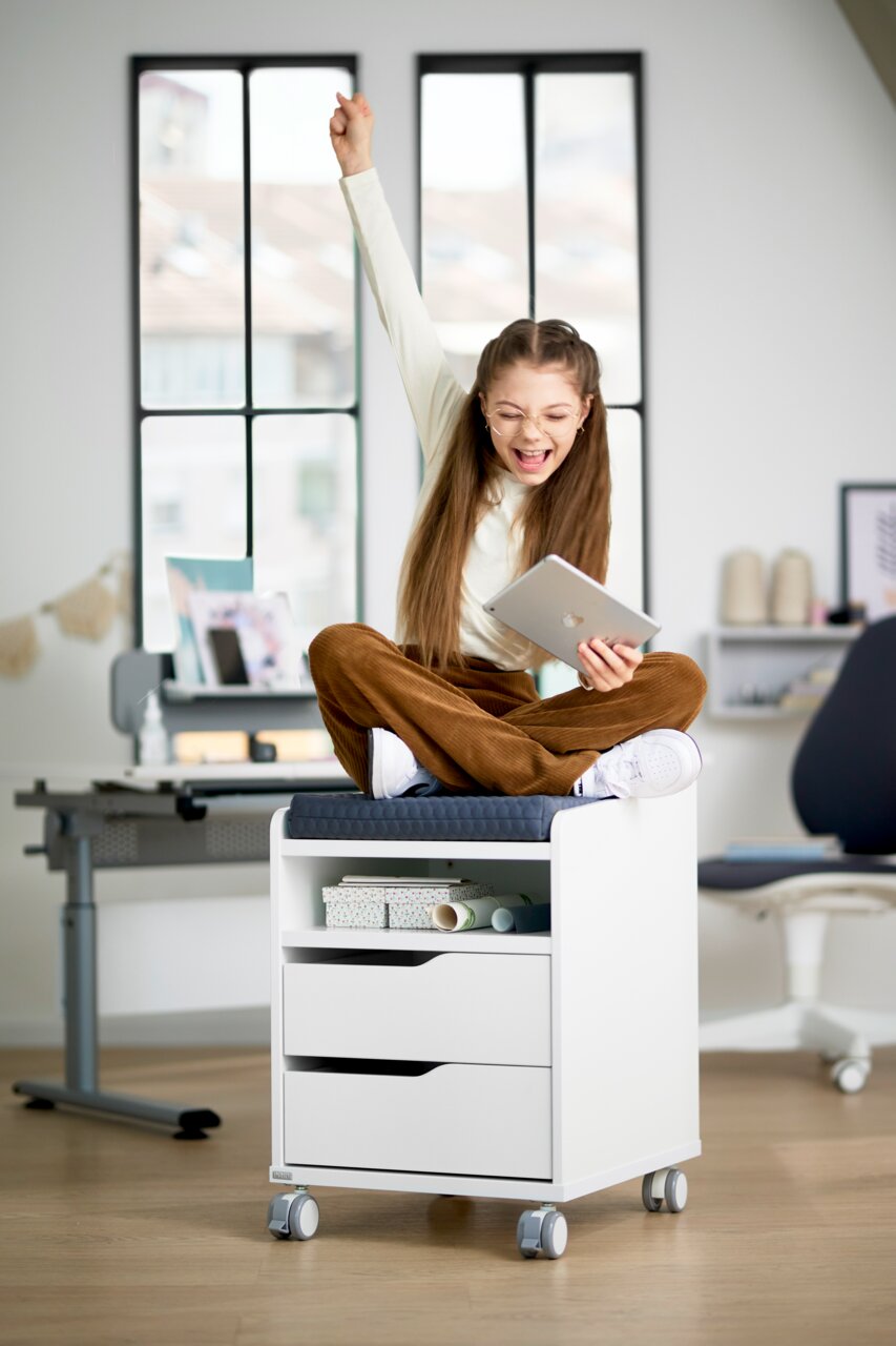 Mädchen sitzt fröhlich auf einem weißen Rollcontainer mit Sitzpolster, Vorderansicht