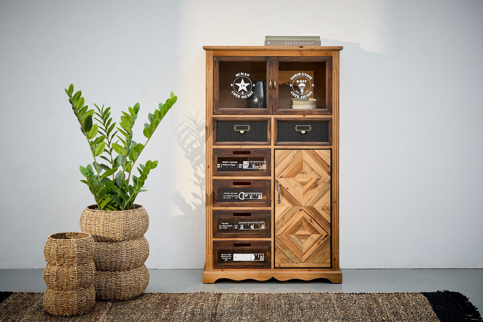 Landscape Kommode ROSITA Kommode aus Holz mit verschiedenen Schubladen und Türen, dekoriert mit Aufdrucken und Mustern, frontal fotografiert. Daneben steht eine Pflanze in einem geflochtenen Korb.