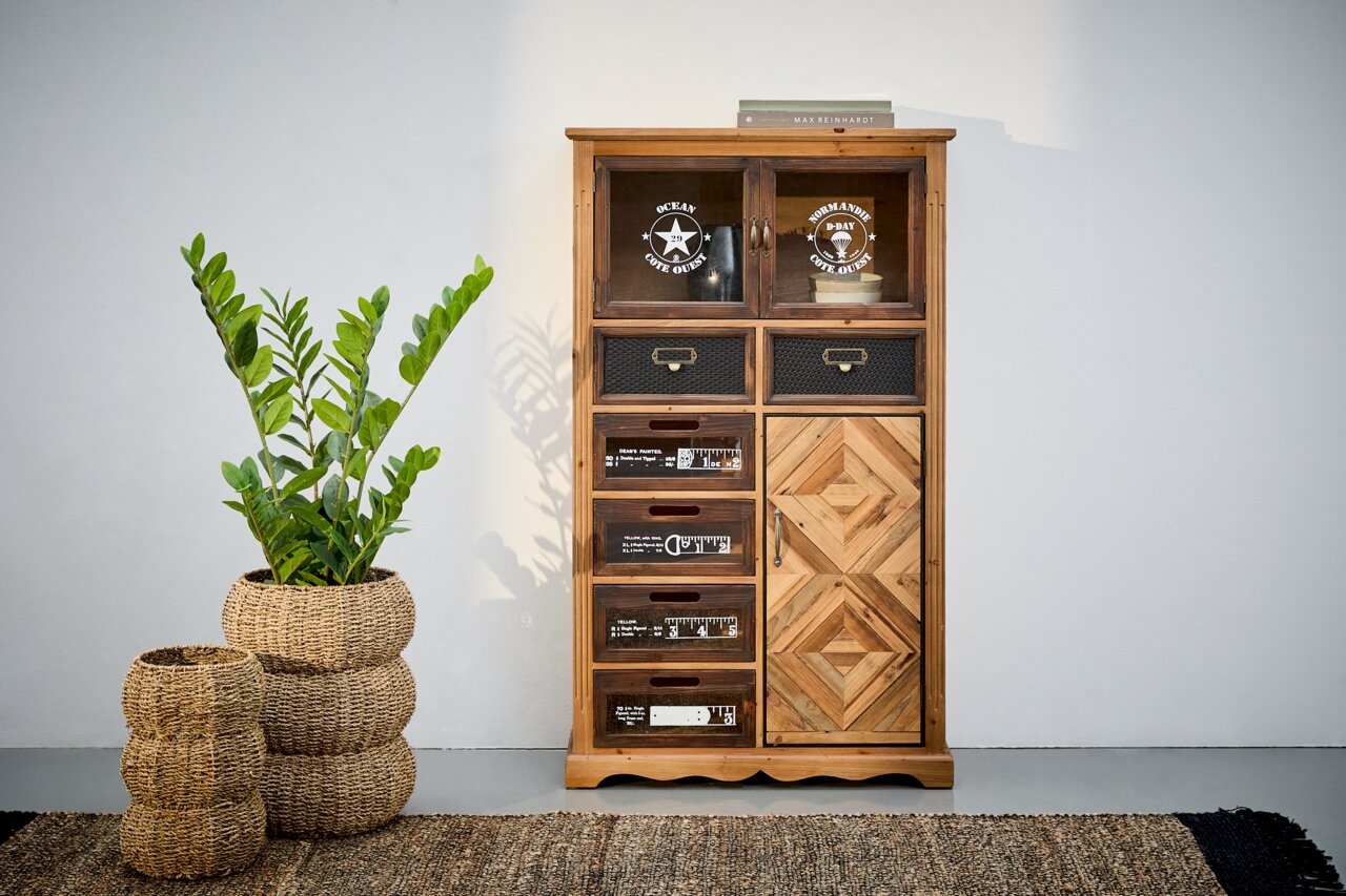 Landscape Kommode ROSITA Kommode aus Holz mit verschiedenen Schubladen und Türen, dekoriert mit Aufdrucken und Mustern, frontal fotografiert. Daneben steht eine Pflanze in einem geflochtenen Korb.