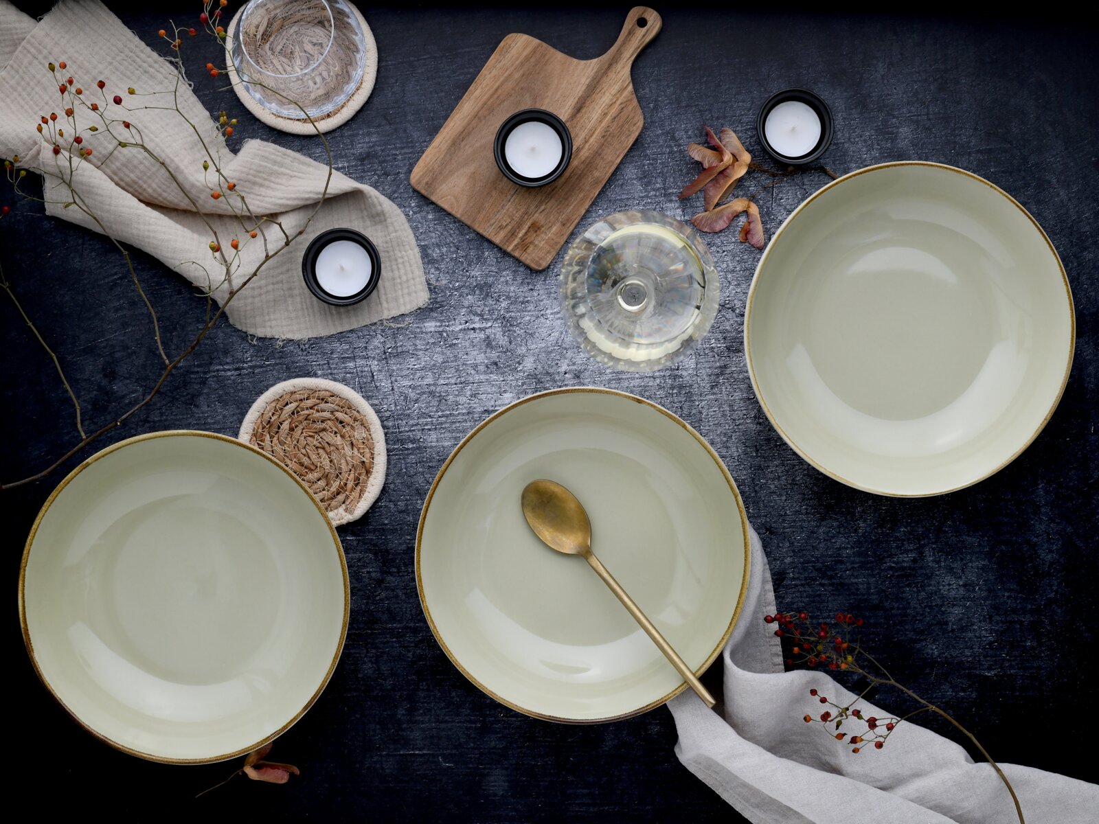 Drei große, hellgrüne Keramikschalen mit braunem Rand auf dunklem Tisch, dekoriert mit Holzbrett, Teelichtern, Glas und Stoffservietten; Draufsicht von oben