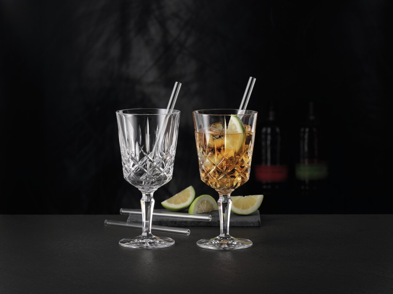 Zwei elegante Cocktailgläser mit Strohhalmen, eines leer und eines mit Getränk und Limettenscheibe, auf schwarzem Hintergrund, seitliche Perspektive.