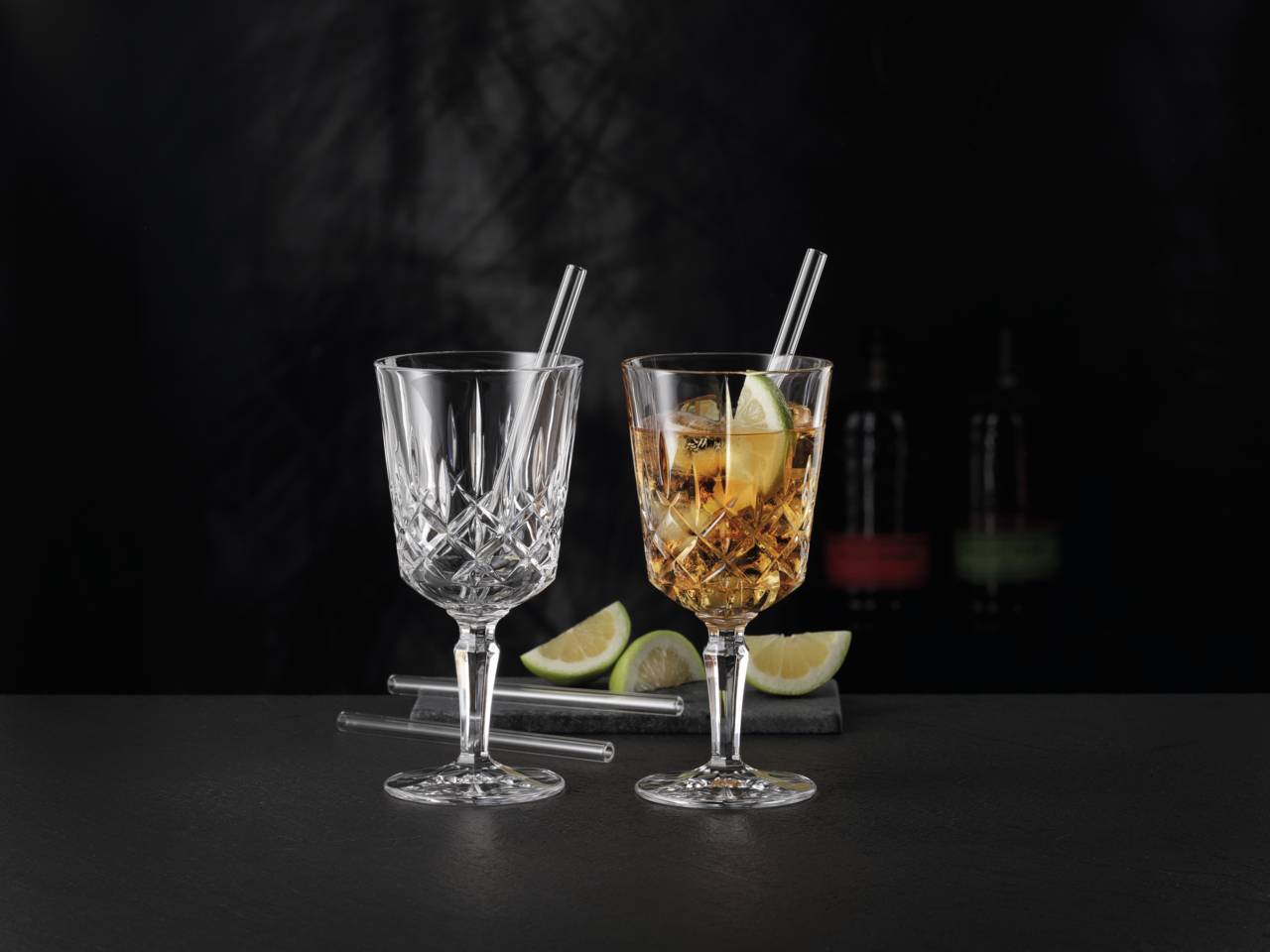 Zwei elegante Cocktailgläser mit Strohhalmen, eines leer und eines mit Getränk und Limettenscheibe, auf schwarzem Hintergrund, seitliche Perspektive.