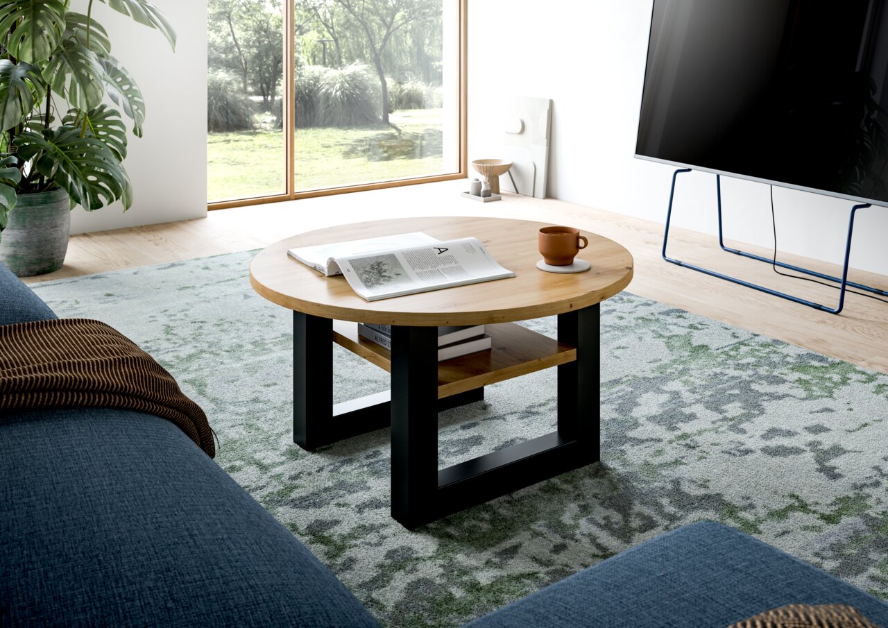 Runder Couchtisch aus Holz mit schwarzem Metallgestell, von schräg oben fotografiert, auf einem gemusterten Teppich in einem modernen Wohnzimmer.