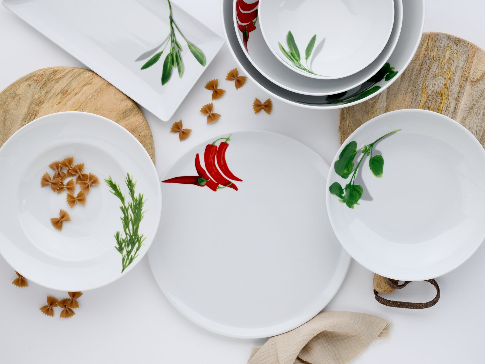 Weißes Porzellan-Geschirrset mit Kräuter- und Chili-Motiven (Teller, Schalen, quadratische Platte) auf weißem Hintergrund mit Holzbrettchen und Pasta, Draufsicht
