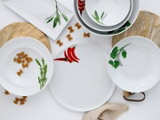 Weißes Porzellan-Geschirrset mit Kräuter- und Chili-Motiven (Teller, Schalen, quadratische Platte) auf weißem Hintergrund mit Holzbrettchen und Pasta, Draufsicht