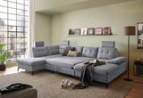 Graues Ecksofa DURBAN in U-Form mit verstellbaren Kopfstützen, dekoriert mit Kissen und Decke, in einem modernen Wohnzimmer mit großen Fenstern und Stadtblick, seitliche Perspektive.