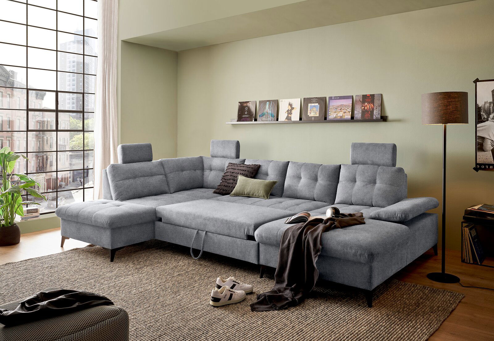 Graues Ecksofa DURBAN in U-Form mit verstellbaren Kopfstützen, dekoriert mit Kissen und Decke, in einem modernen Wohnzimmer mit großen Fenstern und Stadtblick, seitliche Perspektive.