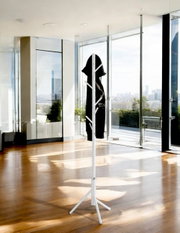 Weißer freistehender Kleiderständer/Garderobenständer mit mehreren Haken und dreibeinigem Standfuß, daran ein schwarzes Kleidungsstück, im hellen Raum mit großen Glasfronten; Perspektive: frontale Ansicht auf Augenhöhe.