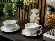 Zwei weiße Kaffeetassen mit Untertassen auf dunkler Küchenarbeitsplatte, eine Tasse mit Cappuccino-Latte-Art, daneben Glasbehälter mit Baiser und Keksen sowie Blumen im Hintergrund, aufgenommen aus leicht erhöhter Frontperspektive.