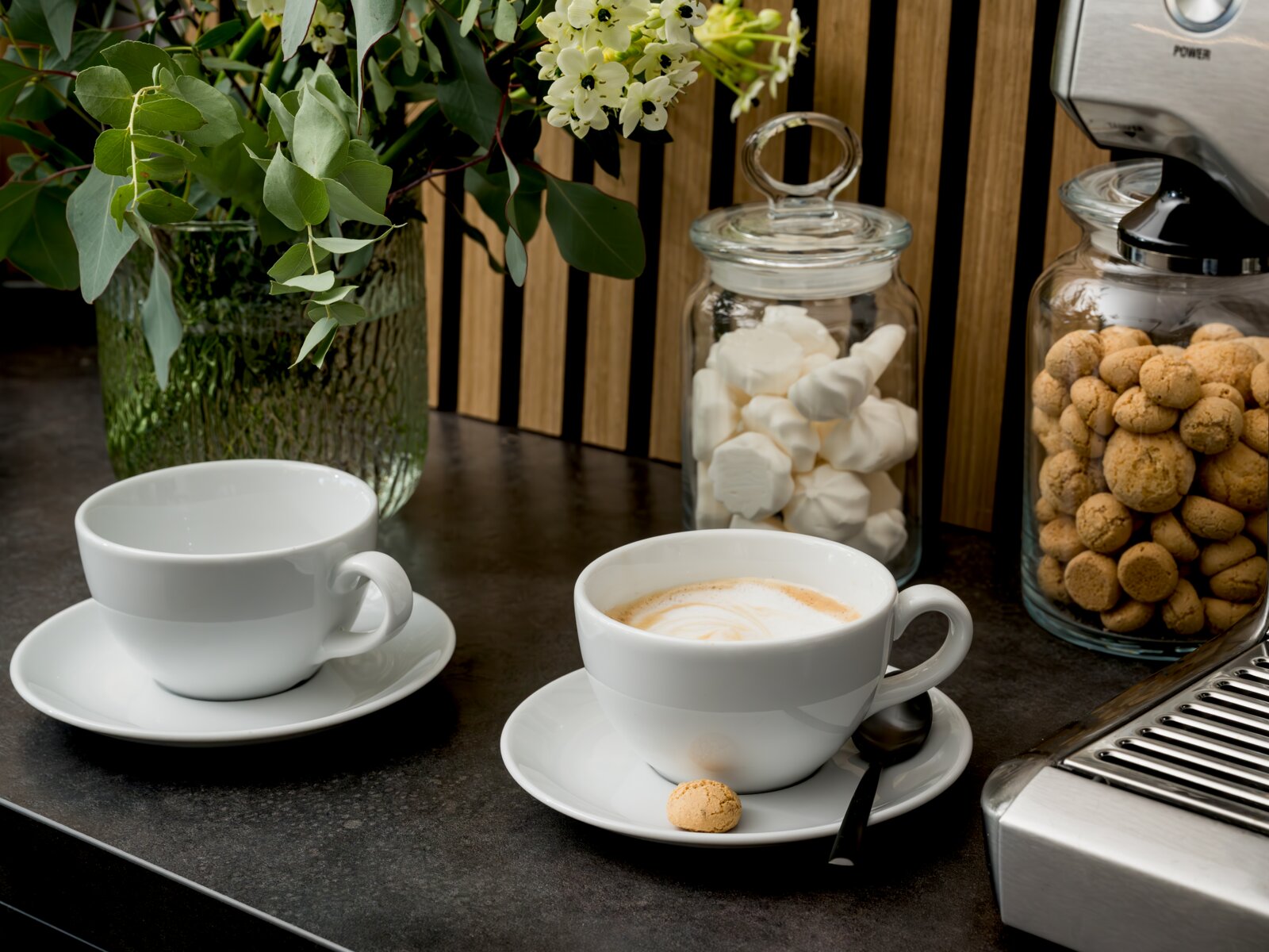 Zwei weiße Kaffeetassen mit Untertassen auf dunkler Küchenarbeitsplatte, eine Tasse mit Cappuccino-Latte-Art, daneben Glasbehälter mit Baiser und Keksen sowie Blumen im Hintergrund, aufgenommen aus leicht erhöhter Frontperspektive.