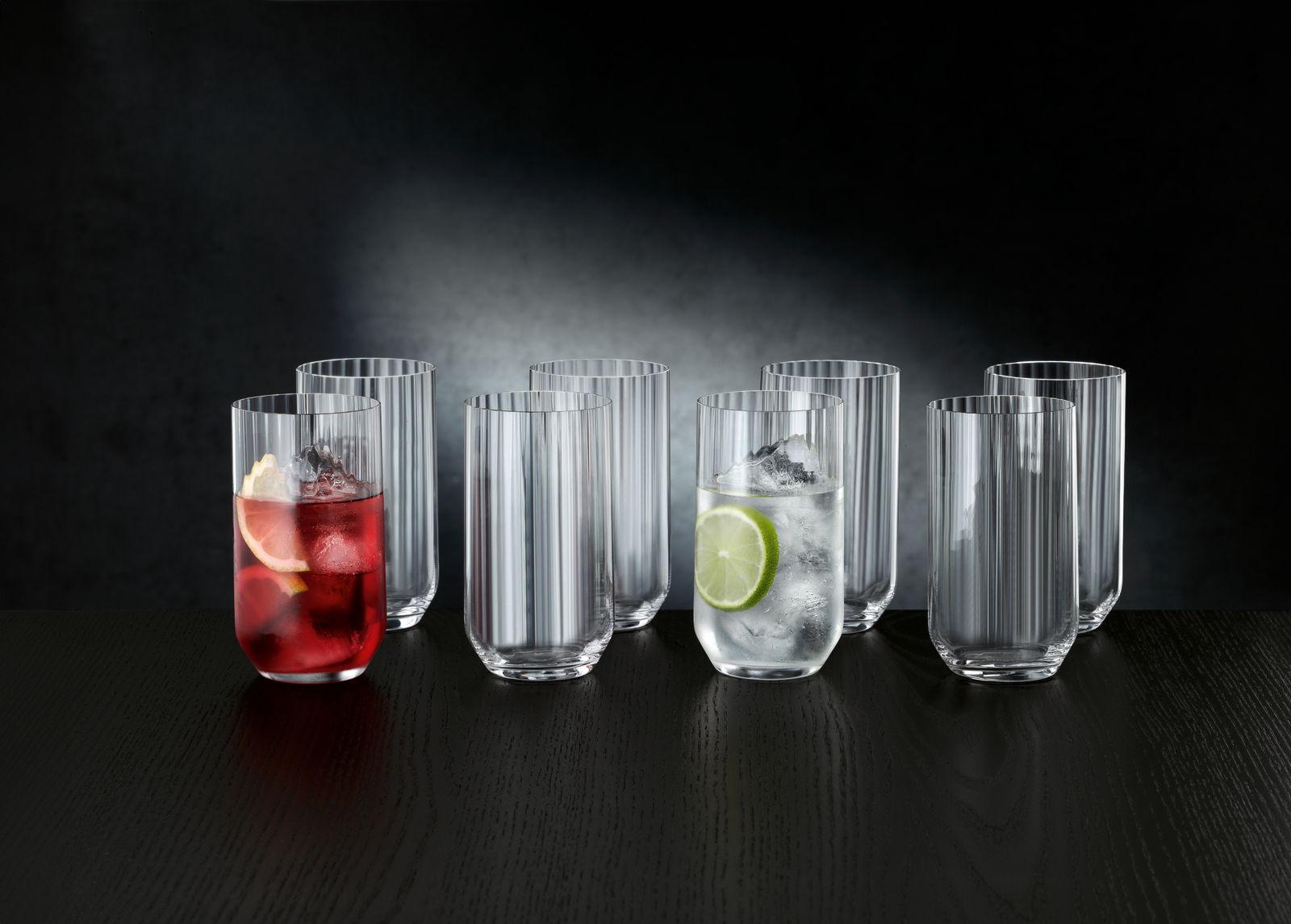 Ein Set aus acht Longdrinkgläsern, teilweise gefüllt mit Getränken und Eis, auf einem dunklen Tisch vor einem dunklen Hintergrund. Die Perspektive ist frontal.