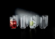Ein Set aus acht Longdrinkgläsern, teilweise gefüllt mit Getränken und Eis, auf einem dunklen Tisch vor einem dunklen Hintergrund. Die Perspektive ist frontal.