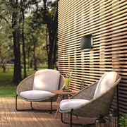 Moderne AKKU-Wandleuchte an einer Holzlattenwand auf einer Terrasse mit zwei gemütlichen Sesseln und einem kleinen Tisch, aus der Perspektive von schräg vorne.