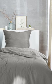 Graue Bettwäsche im Stone-Washed-Look auf einem Bett, fotografiert aus der Vorderansicht, mit dekorativen Elementen im Hintergrund.