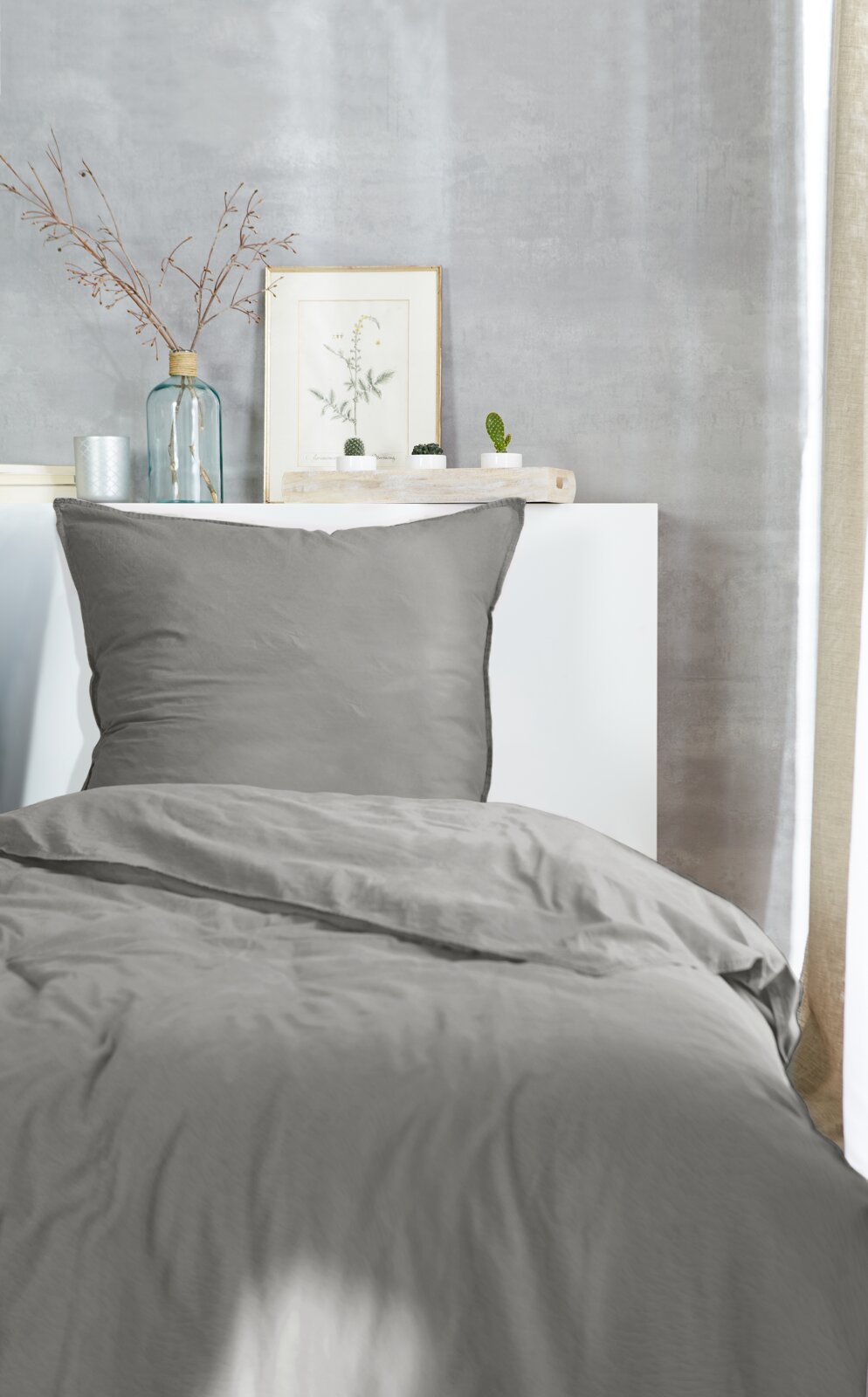 Graue Bettwäsche im Stone-Washed-Look auf einem Bett, fotografiert aus der Vorderansicht, mit dekorativen Elementen im Hintergrund.