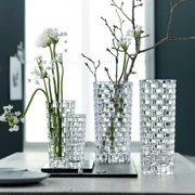 Drei elegante, durchsichtige Vasen mit geometrischem Muster, gefüllt mit weißen Blumen und Zweigen, auf einem Tisch vor einem Fenster.