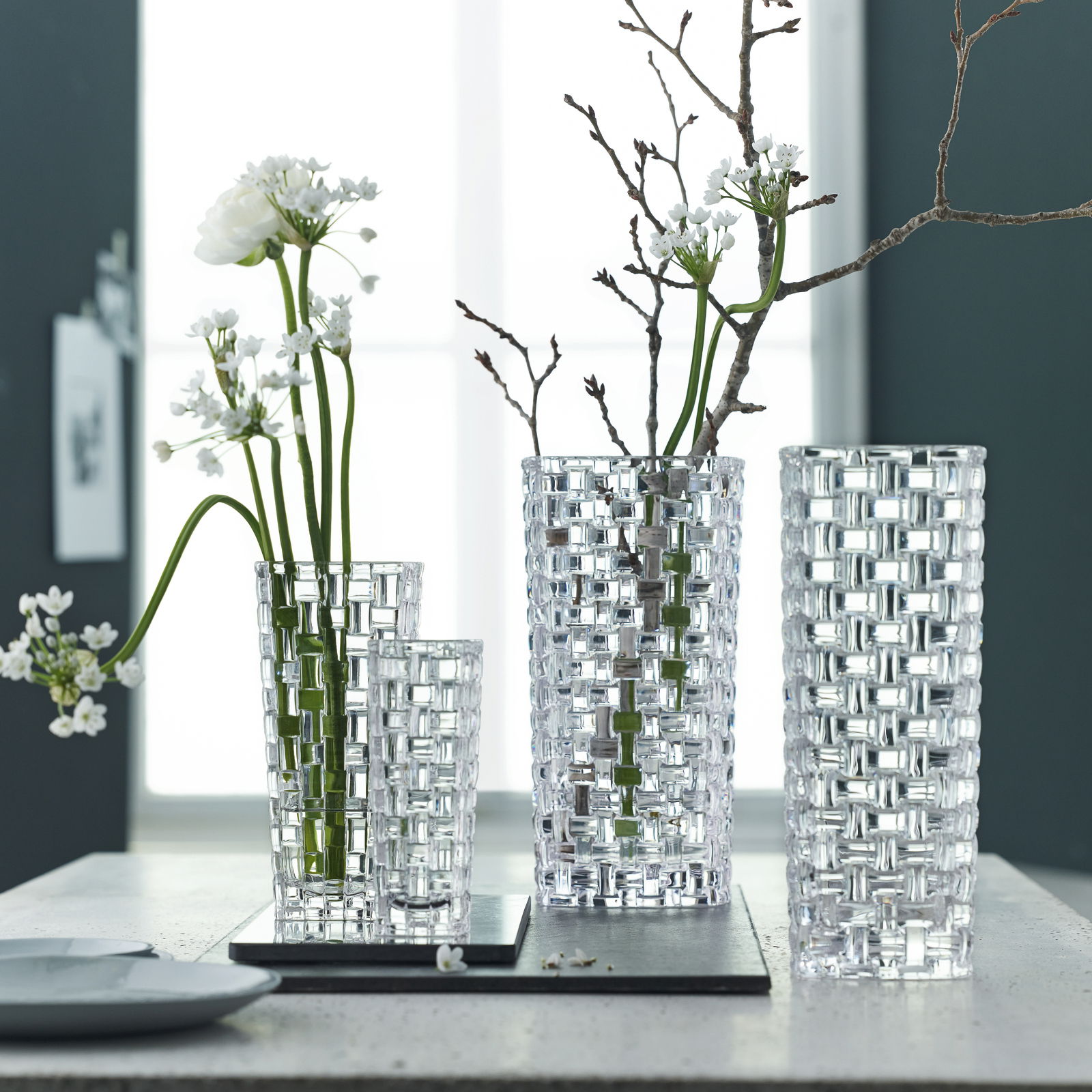 Drei elegante, durchsichtige Vasen mit geometrischem Muster, gefüllt mit weißen Blumen und Zweigen, auf einem Tisch vor einem Fenster.