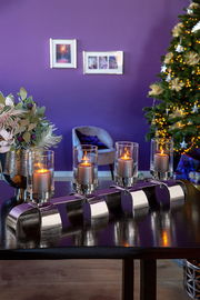 Moderner vierflammiger Kerzenleuchter mit Glaszylindern auf einem Tisch, im Hintergrund ein festlich geschmückter Raum mit Weihnachtsbaum, seitliche Perspektive.