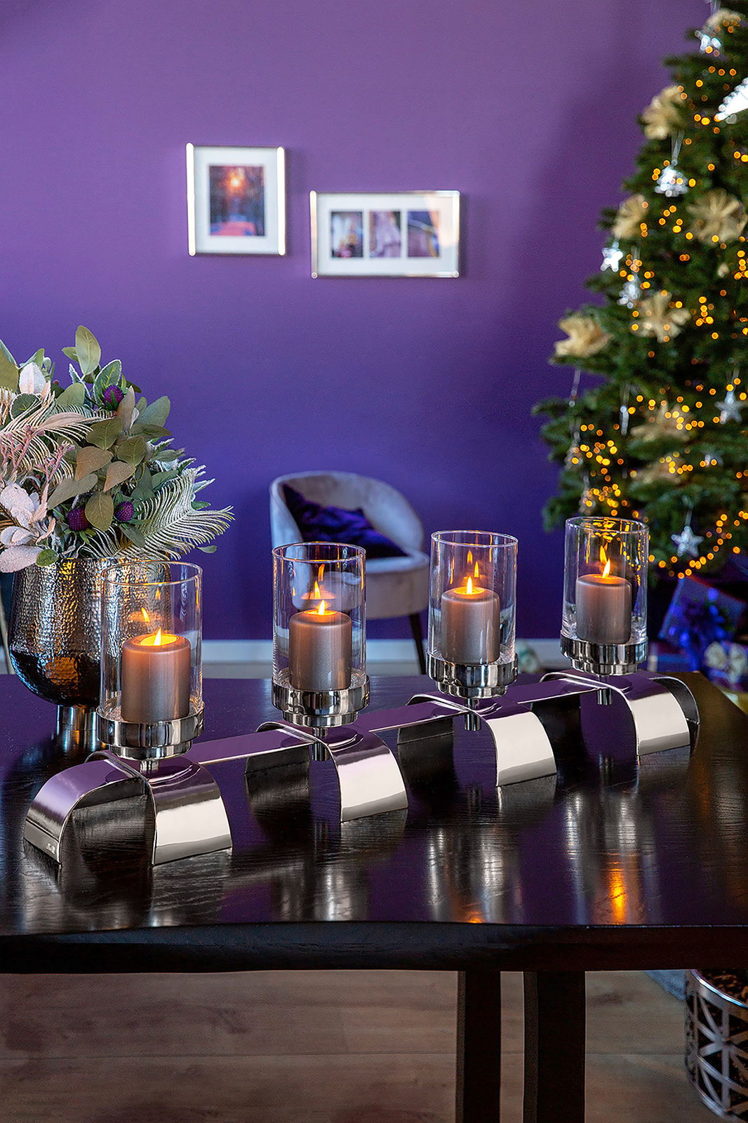 Moderner vierflammiger Kerzenleuchter mit Glaszylindern auf einem Tisch, im Hintergrund ein festlich geschmückter Raum mit Weihnachtsbaum, seitliche Perspektive.