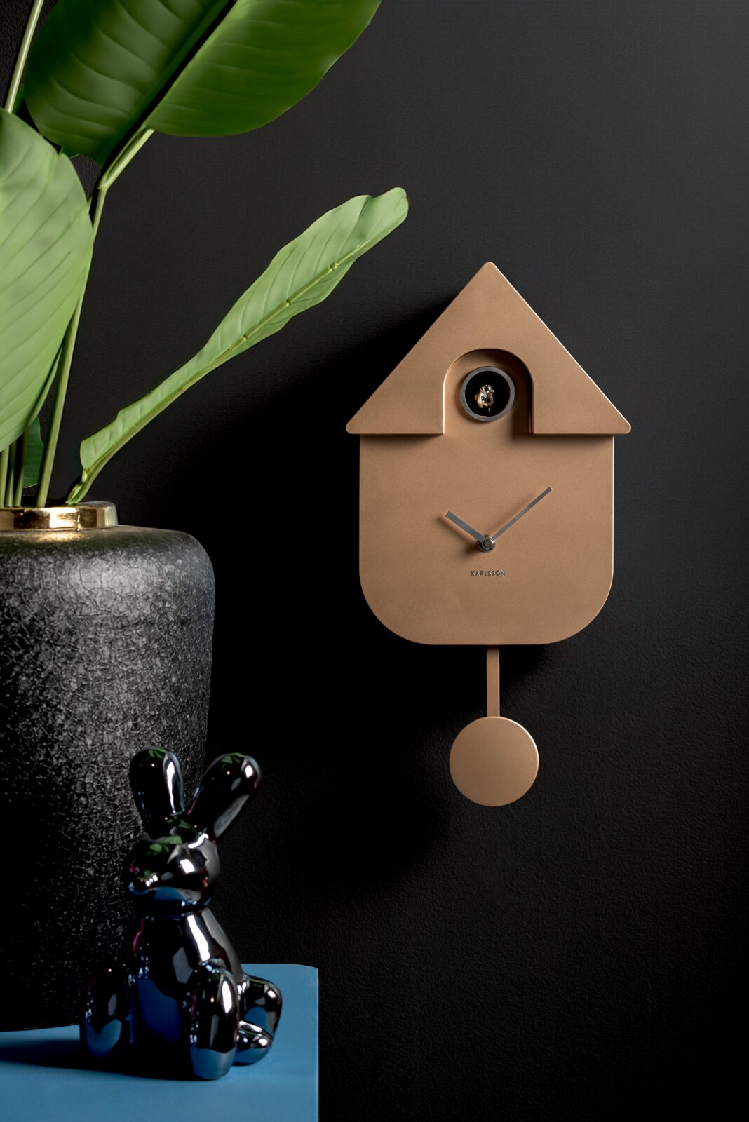 Moderne Wanduhr in Form eines Kuckucks mit Pendel, in einem eleganten Raum mit Pflanzen und Dekorationen, seitliche Perspektive.