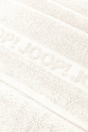 Nahaufnahme in schräger Draufsicht auf ein weißes, flauschiges Handtuch mit eingewebtem/erhabenem Schriftzug „JOOP!“.