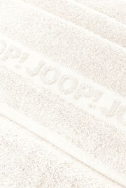 Nahaufnahme in schräger Draufsicht auf ein weißes, flauschiges Handtuch mit eingewebtem/erhabenem Schriftzug „JOOP!“.