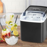 Schwarzer Eiswürfelbereiter mit transparentem Deckel, gefüllt mit Eiswürfeln, auf einer Küchenarbeitsplatte. Im Vordergrund stehen Gläser mit Getränken und Eiswürfeln. Perspektive von vorne rechts.