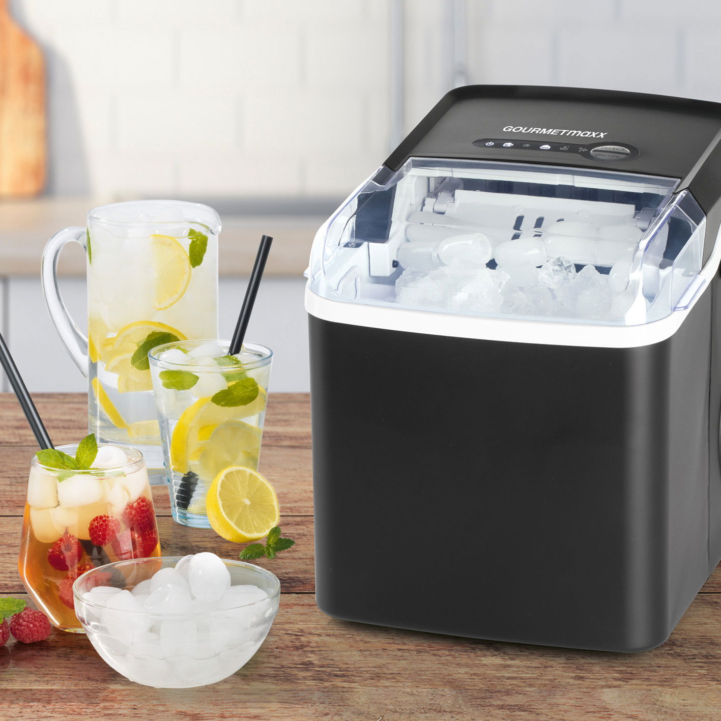 Schwarzer Eiswürfelbereiter mit transparentem Deckel, gefüllt mit Eiswürfeln, auf einer Küchenarbeitsplatte. Im Vordergrund stehen Gläser mit Getränken und Eiswürfeln. Perspektive von vorne rechts.