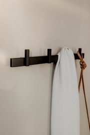Schwarze, wandmontierte Garderobenleiste mit zwei Haken, daran hängt ein weißes Tuch und ein brauner Lederriemen; seitliche Perspektive an heller Wand