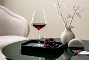 Ein elegantes Burgunderkelch-Glas, gefüllt mit Rotwein, steht auf einem Tablett neben einer Traube auf einem grünen Tisch. Im Hintergrund ist eine Vase mit dekorativen Zweigen zu sehen. Perspektive: seitlich.