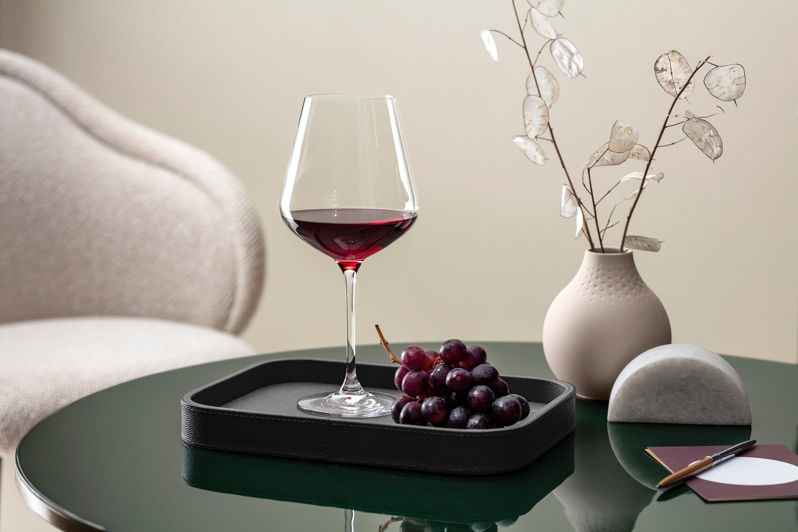 Villeroy & Boch Set van 4 Bourgondische wijnglazen LA DAVINA Ein elegantes Burgunderkelch-Glas, gefüllt mit Rotwein, steht auf einem Tablett neben einer Traube auf einem grünen Tisch. Im Hintergrund ist eine Vase mit dekorativen Zweigen zu sehen. Perspektive: seitlich.