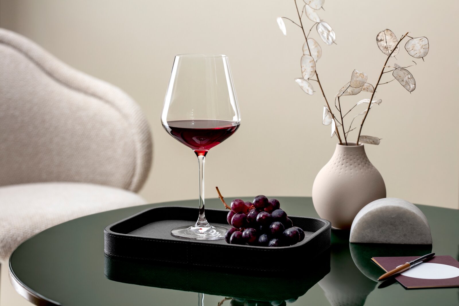 Een elegant bordeauxglas, gevuld met rode wijn, staat op een dienblad naast een tros druiven op een groene tafel. Op de achtergrond is een vaas met decoratieve takjes te zien. Perspectief: zijaanzicht.