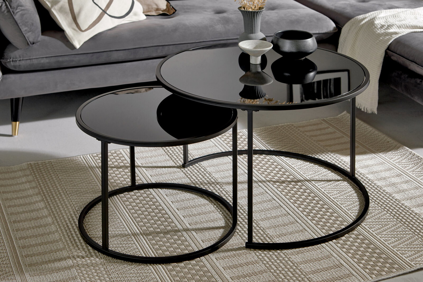 Zweier-Set Couchtische mit schwarzer Glasplatte und Metallgestell, perspektivisch von vorne fotografiert, auf einem gemusterten Teppich vor einem grauen Sofa.