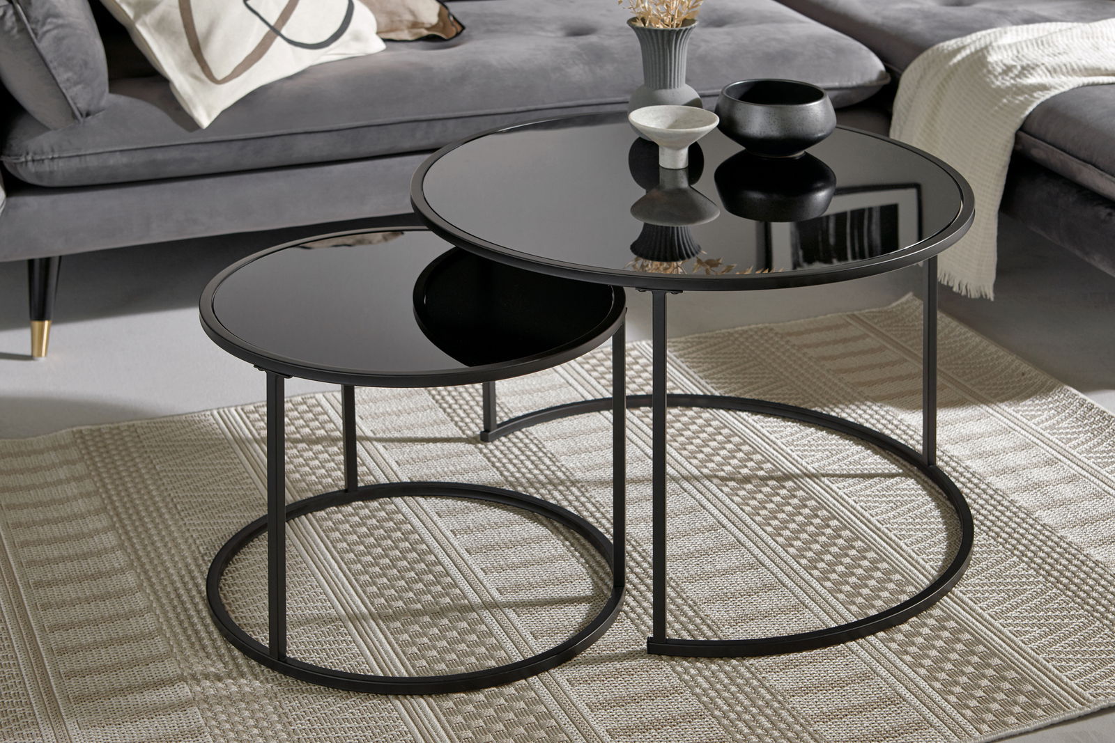 Zweier-Set Couchtische mit schwarzer Glasplatte und Metallgestell, perspektivisch von vorne fotografiert, auf einem gemusterten Teppich vor einem grauen Sofa.