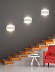 Moderne Wandleuchte Concha in minimalistischem Design, montiert an einer grauen Wand über einer Treppe, mit warmem Licht nach oben und unten strahlend.
