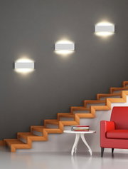 Moderne wandlamp Concha in minimalistisch design, bevestigd aan een grijze muur boven een trap, met warm licht dat zowel naar boven als naar beneden schijnt.